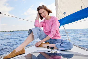 A woman wears a bright pink sweater paired with light wash embroidered jeans while sitting on a boat.