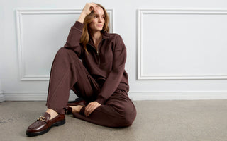 A woman wearing a brown zip-neck sweatshirt and matching sweatpants, seated on the floor.