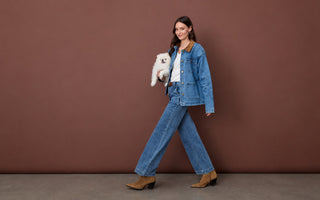 A woman wearing a denim jacket and matching wide-leg jeans, holding a small white dog.