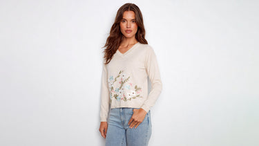 A woman wearing a beige V-neck sweater with floral embroidery and light wash denim jeans.