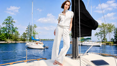 A woman wearing a light grey zip-neck top and matching wide-leg pants, standing on a sailboat near a marina.