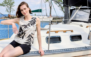 Women's sleeveless linen tank top with black tropical palm leaf print on natural beige background, styled with black biker shorts, worn on a sailboat by the water.