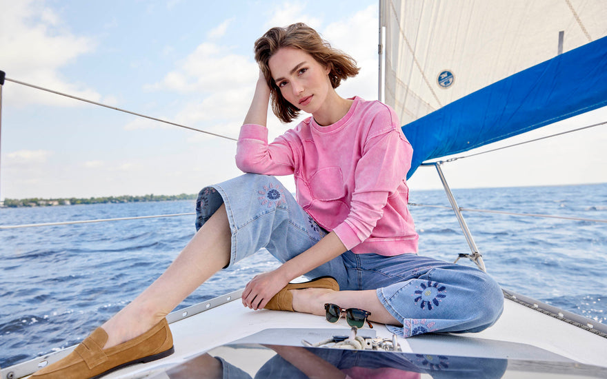 A woman wears a bright pink sweater paired with light wash embroidered jeans while sitting on a boat.