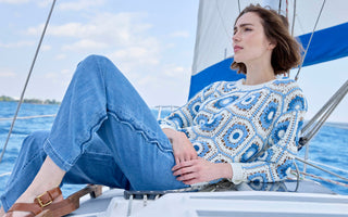 A woman wearing a blue and white geometric crochet sweater with wide leg denim pants, reclining on a sailboat.