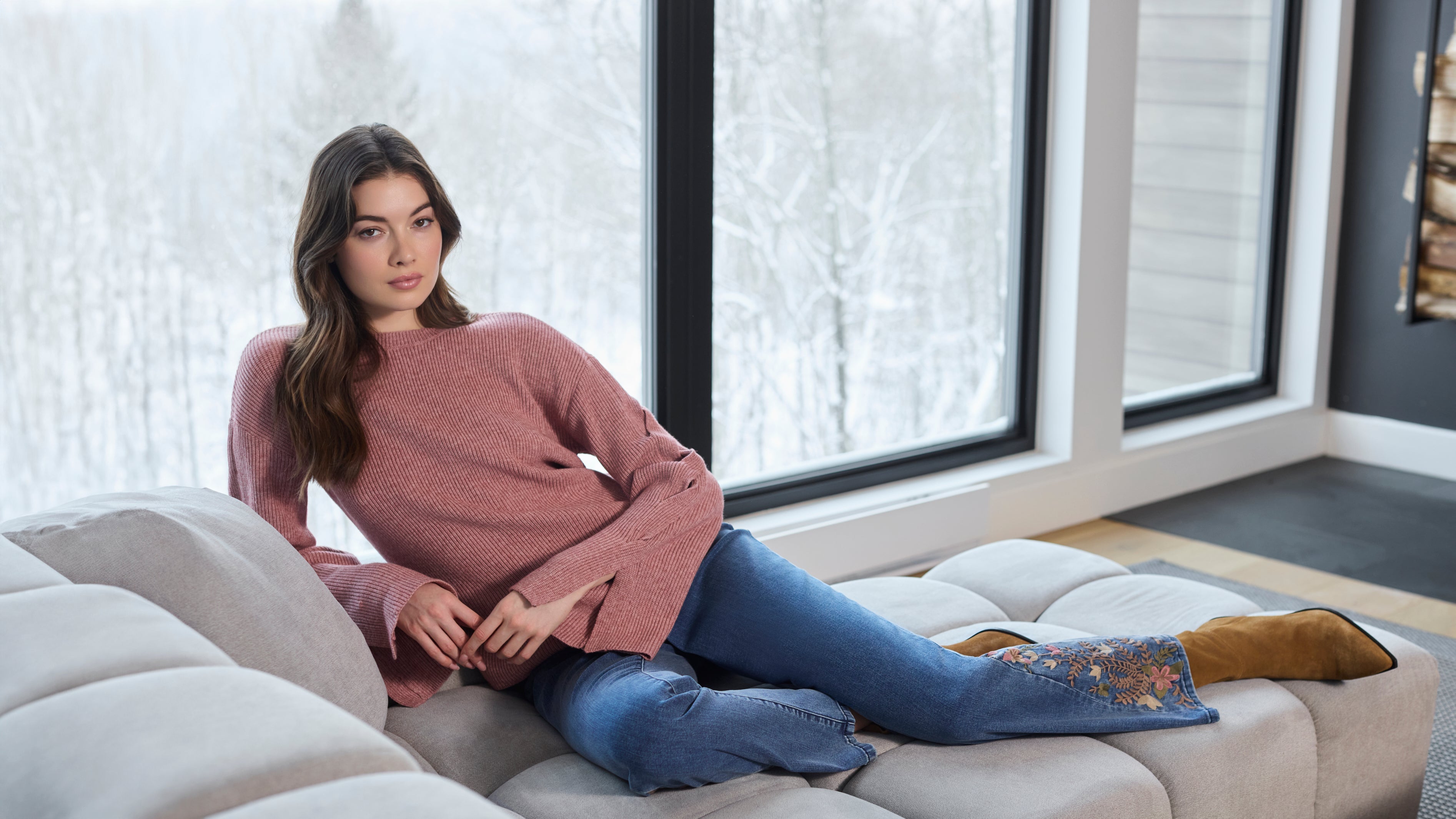 Woman lounging on a light-gray sectional sofa, wearing a pink long-sleeve sweater, blue embroidered jeans, and brown boots, with a snowy winter landscape visible through large windows.