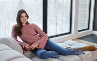 Woman lounging on a light-gray sectional sofa, wearing a pink long-sleeve sweater, blue embroidered jeans, and brown boots, with a snowy winter landscape visible through large windows.
