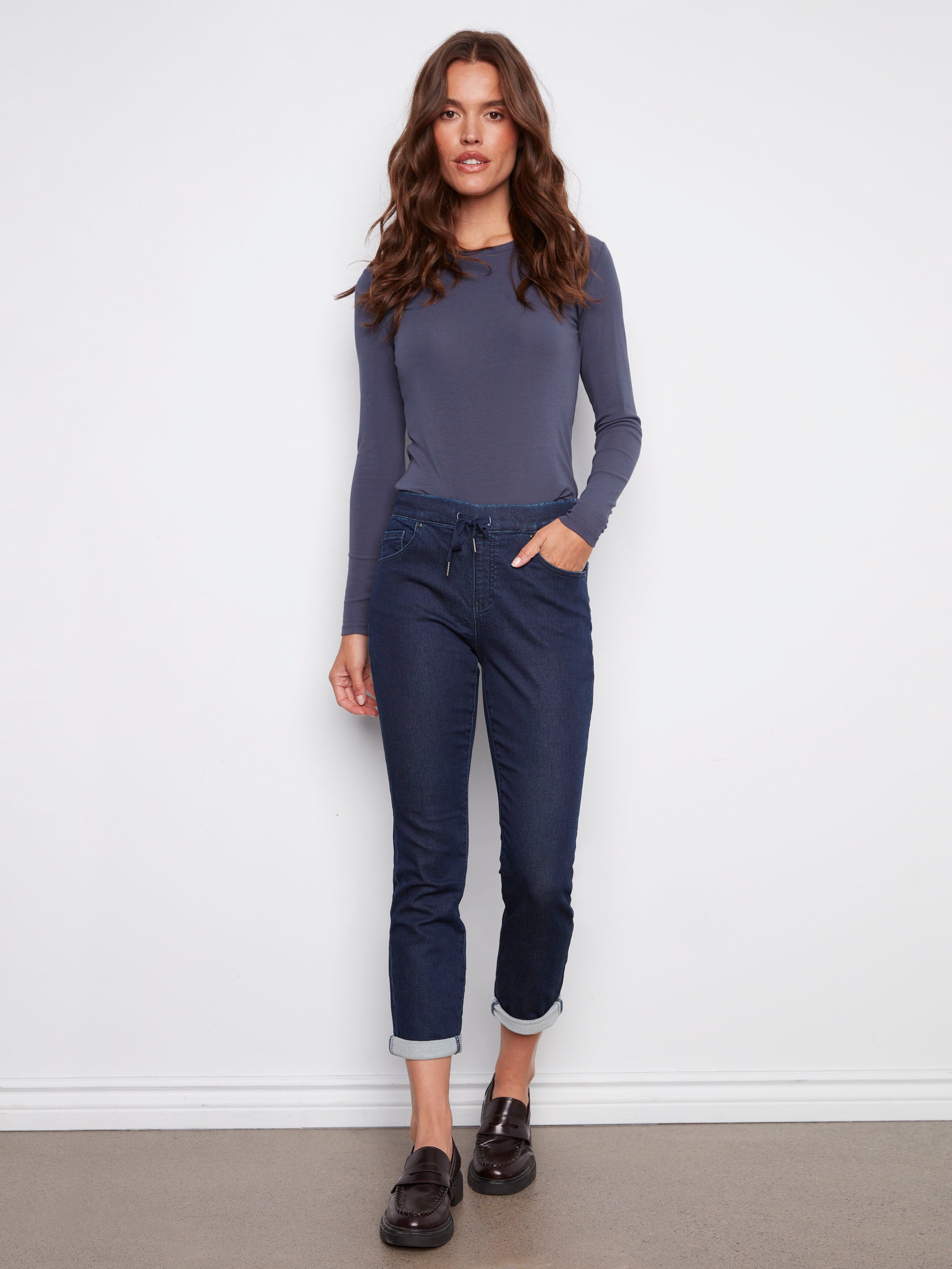 Model wearing dark blue jogger jeans with drawstring waist, paired with a fitted long-sleeve slate blue top and black loafers, standing against white wall