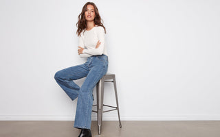 Woman sitting on a stool wearing a white sweater and blue wide leg jeans against a plain background
