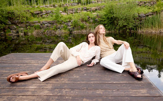 Two women on a wooden dock wearing Charlie B ivory embroidered blouse and beige striped knit top with cream linen pants.