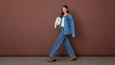 A woman wearing a denim jacket and matching wide-leg jeans, holding a small white dog.