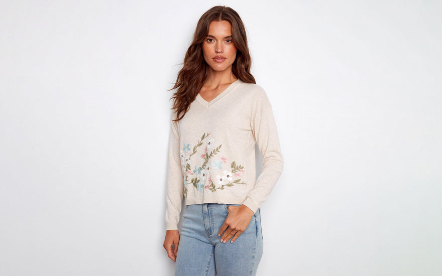 A woman wearing a beige V-neck sweater with floral embroidery and light wash denim jeans.
