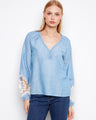 Blue blouse featuring a braided V-neck design, paired with jeans for a stylish look. By Charlie B.